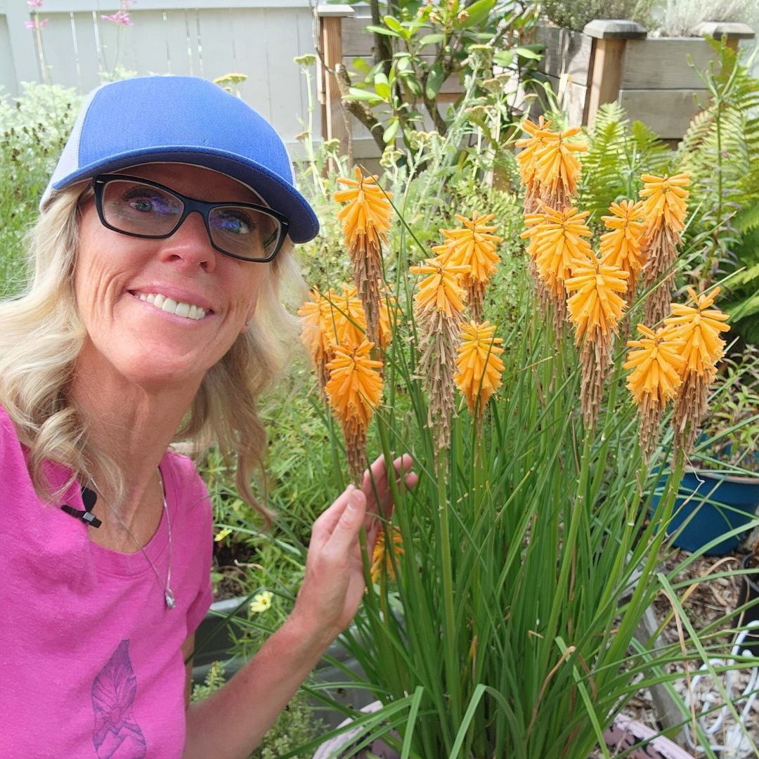 Close up of a woman with blonde hair wearing glasses and a blue hat next to grassy greens with yellow-orange blooms