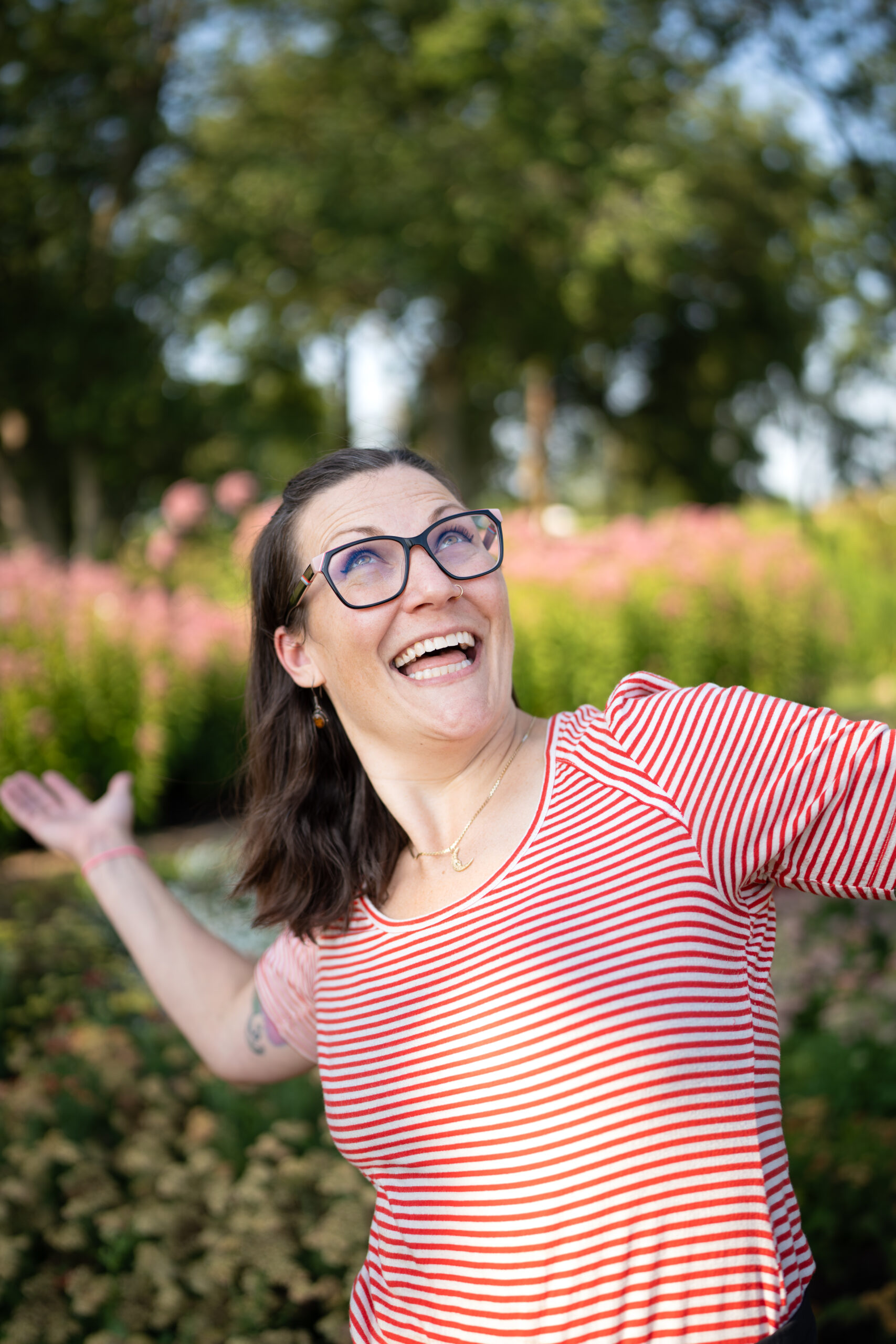 Smiling woman with brown hair, wearing glasses and a red striped shirt posing with her arms out in excitement in front of a blurred field of flowers