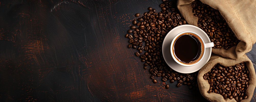 bird's eye view of a white cup filled with coffee sitting on a white saucer surrounded by 2 burlap bags of coffee beans spilling out onto a dark wooden surface
