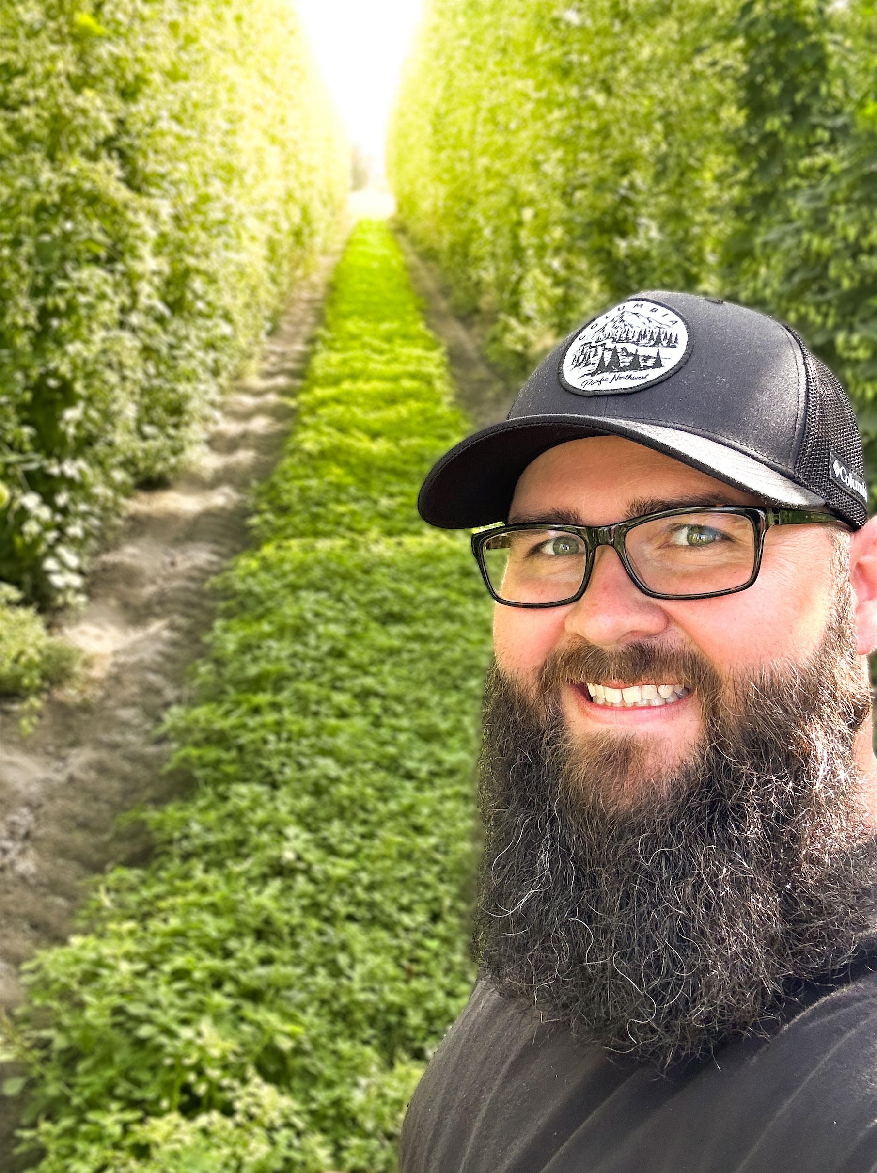 Headshot of a man with dark beard, glasses, and black cap standing in front of lush green walkway