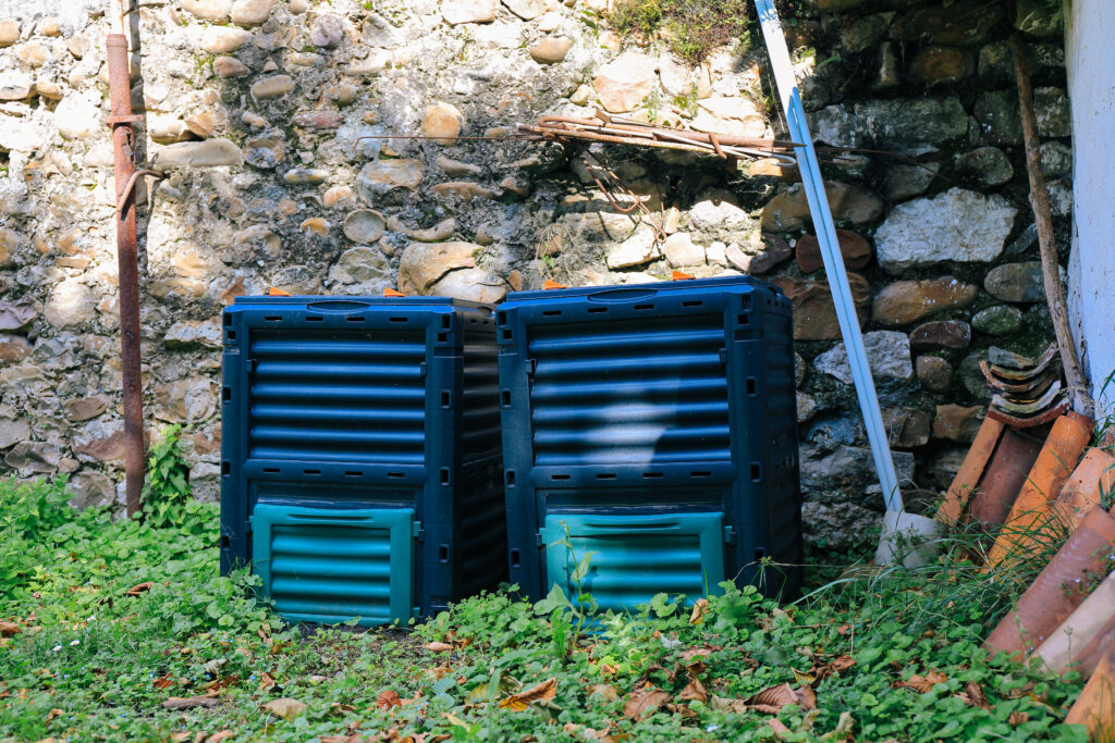 Two dark blue plastic compost bins sitting on grass against a stone wall