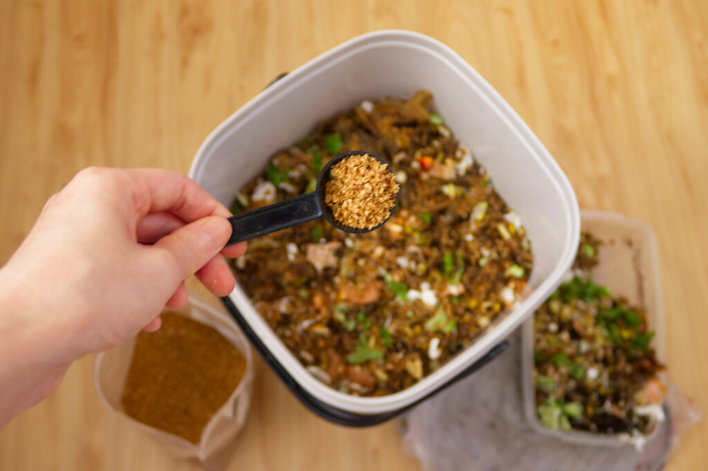 A hand holding a tablespoon of Bokashi bran over a container of food scraps
