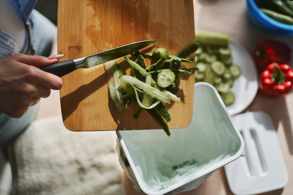 A kitchen knife scraping food scraps off a wooden cutting board into a white container; you can see blurred food prep on a counter in the background