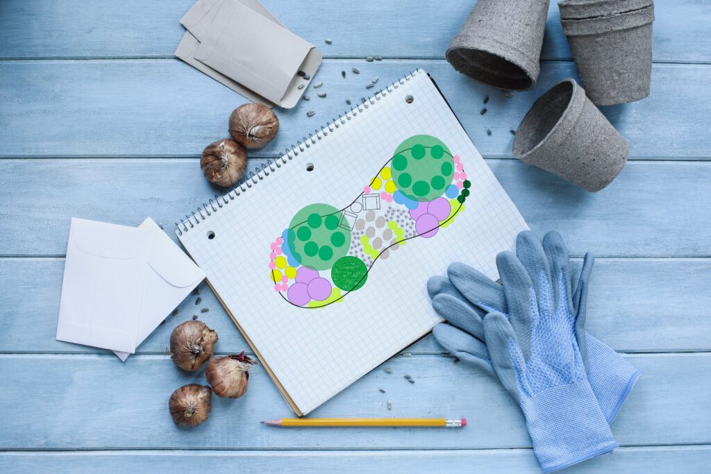 Gardening journal grid notebook with flower bed plan surrounded by garden gloves, pencil, seeds, flower bulbs, envelopes and peat pots on a rustic wooden table. Table top view. 